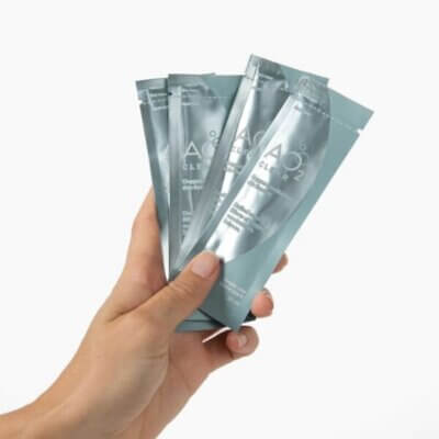 Hand holding several silver skincare sachets labeled AOA cleanser, fanned out in the palm.