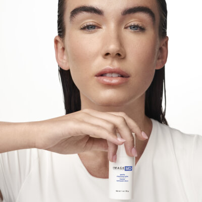 Close-up of a woman in a white top holding a white skincare bottle labeled IMAGE MD, focusing on the product near her chin.