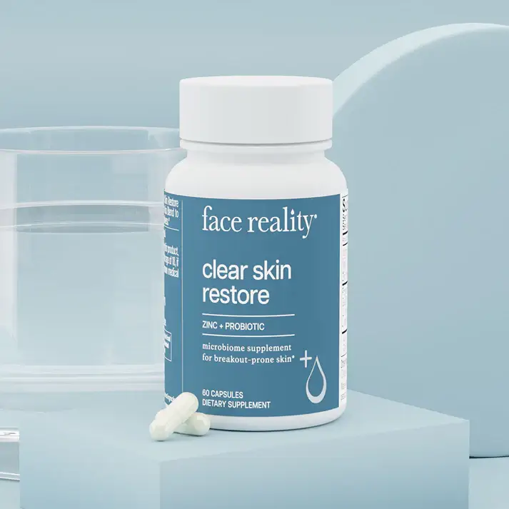 Blue-labeled bottle of face reality clear skin restore dietary supplement with two white capsules beside it on a pale blue display.
